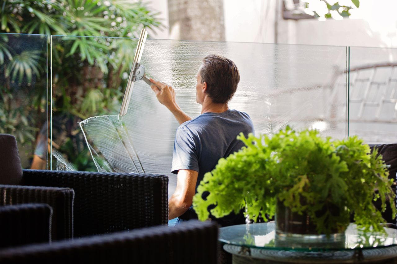services-01 A man cleaning a glass barrier outdoors with a squeegee, surrounded by greenery.